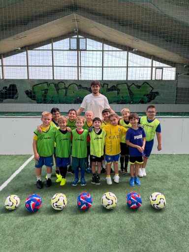 Gruppe von Kindern in Fußballtrikots, zusammen mit einem Trainer, vor einer Sporthalle.
