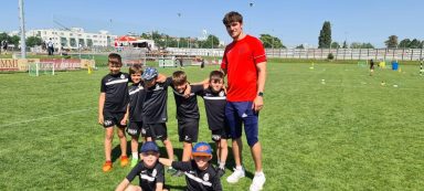 Sechs Kinder in Sportkleidung posieren mit einem Trainer auf einem grünen Rasenfeld.